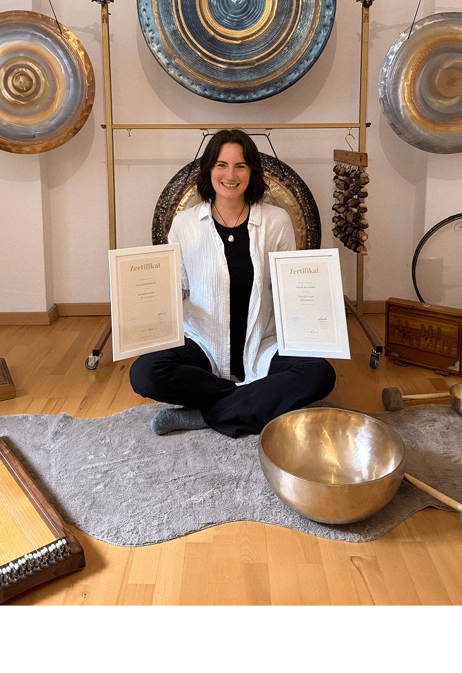 Sarah Remböck sitzt lächelnd mit zwei Zertifikaten zwischen Gongs und einer großen Klangschale